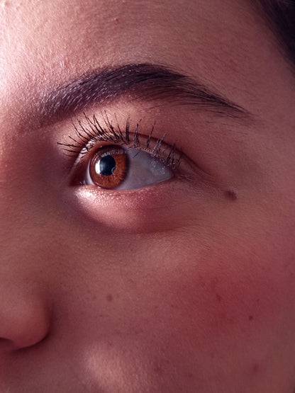 Extreme close-up of a human eye with long dark eyelashes, a brown iris, and a dark eyebrow on warm skin.